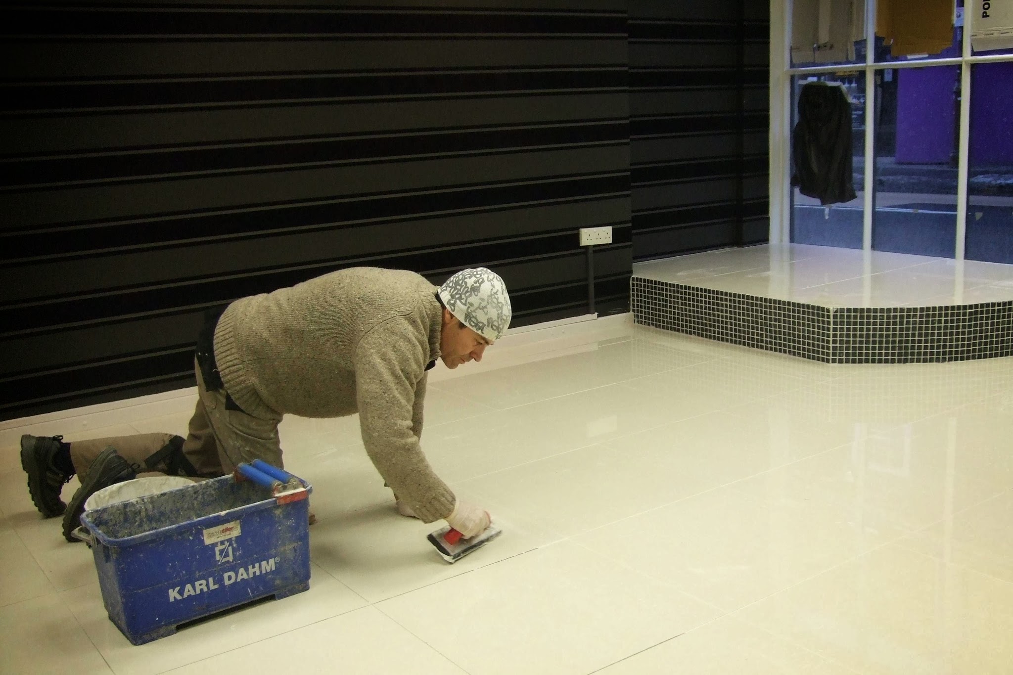 Tiler on his knees grouting a large cream-tiled floor beside a mosaic-faced step
